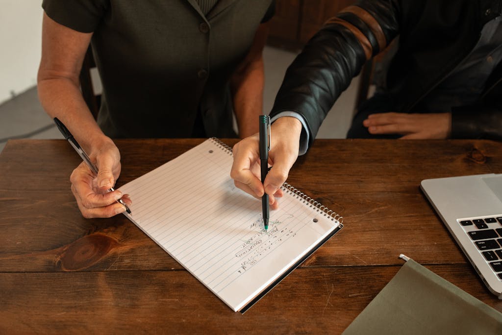 Two people writing notes during a collaborative meeting. Hands, notebook, and pens on wooden table.